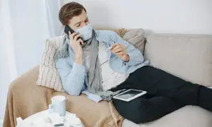 Chory mężczyzna w masce i szaliku leży na kanapie, rozmawiając przez telefon i trzymając termometr.