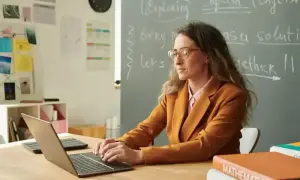 Nauczyciel początkujący z okularami pracuje przy laptopie w klasie. Na tablicy napisy po angielsku.