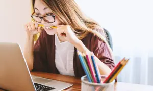 Kobieta w okularach gryzie ołówek, pracując przy laptopie. Kolorowe kredki w pojemniku sugerują kreatywność, ale jej mina zdradza trudności w osiągnięciu work life balance.