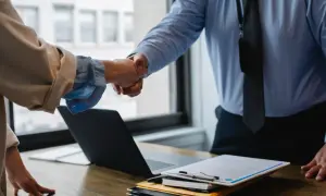 Dwie osoby ściskają sobie dłonie, symbolizując współpracę i inkluzywność w biznesie. Na stole leży laptop i dokumenty.