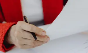 Dłoń w czerwonej marynarce trzyma długopis nad dokumentem, zastanawiając się, czy umowa zlecenie wlicza się do emerytury.