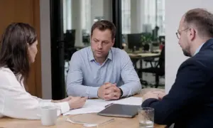 Trzech ludzi przy stole. Mężczyzna w jasnej koszuli wygląda na zmartwionego, być może rozmawia o molestowaniu.