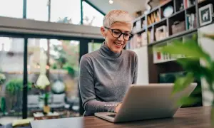 Uśmiechnięta kobieta z krótkimi blond włosami i okularami pracuje na laptopie, szukając informacji, czy na umowę zlecenie należy się dodatek nocny.