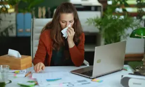 Kobieta z chusteczką w dłoni siedzi przy biurku, zastanawiając się, czy L4 na umowie zlecenie jest płatne. Na stole laptop, dokumenty i pudełko chusteczek.