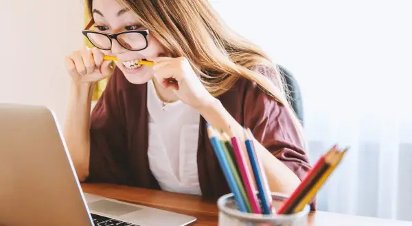Kobieta w okularach gryzie ołówek, pracując przy laptopie. Kolorowe kredki w pojemniku sugerują kreatywność, ale jej mina zdradza trudności w osiągnięciu work life balance.