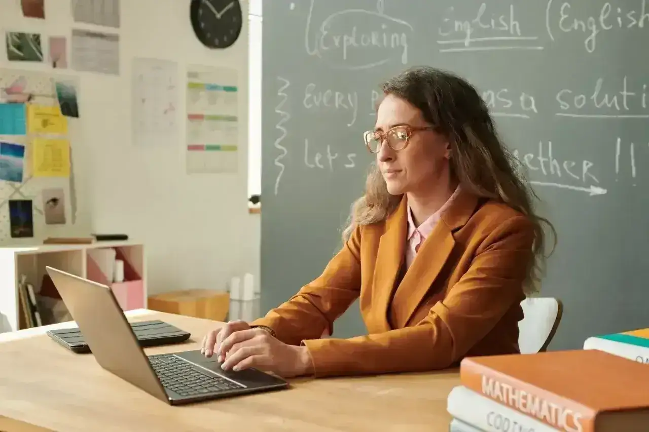 Nauczyciel początkujący z okularami pracuje przy laptopie w klasie. Na tablicy napisy po angielsku.