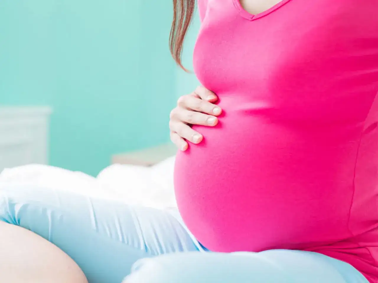 Kobieta w 34. tygodniu ciąży, ubrana w różową bluzkę i niebieskie spodnie, delikatnie dotyka swojego brzucha.