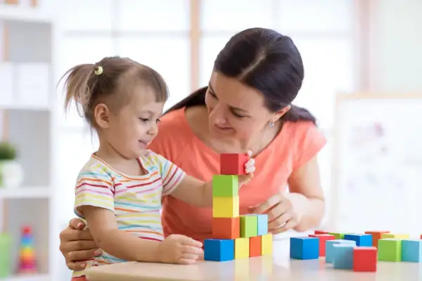 Uśmiechnięta kobieta i dziewczynka budują wieżę z kolorowych klocków. To obrazek, który może inspirować, jak zatrudnić nianię.