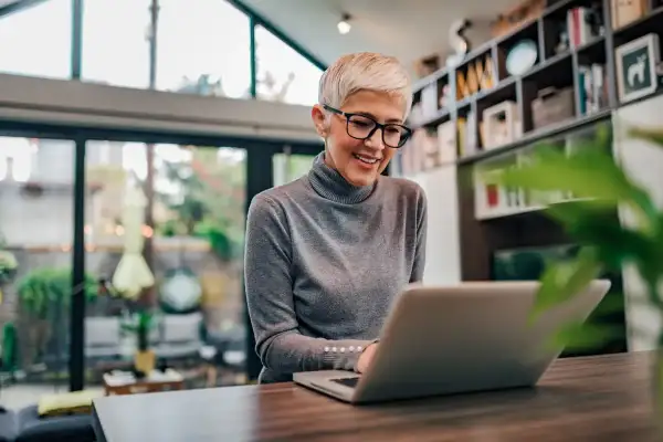 Uśmiechnięta kobieta z krótkimi blond włosami i okularami pracuje na laptopie, szukając informacji, czy na umowę zlecenie należy się dodatek nocny.