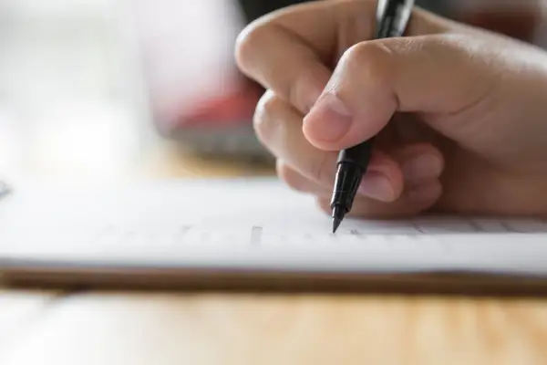 Dłoń pisze piórem po papierze. Czy można skończyć technikum bez egzaminu zawodowego? Zastanawiam się nad tym, wypełniając dokumenty.